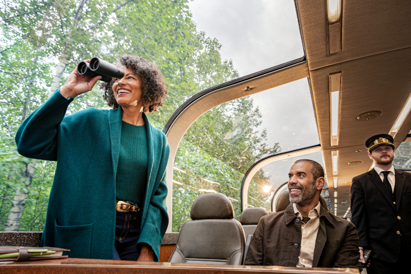 A woman and man travelling in a domed railroad car in Alaska. The woman is holding binoculars. There is a train conductor standing behind them.