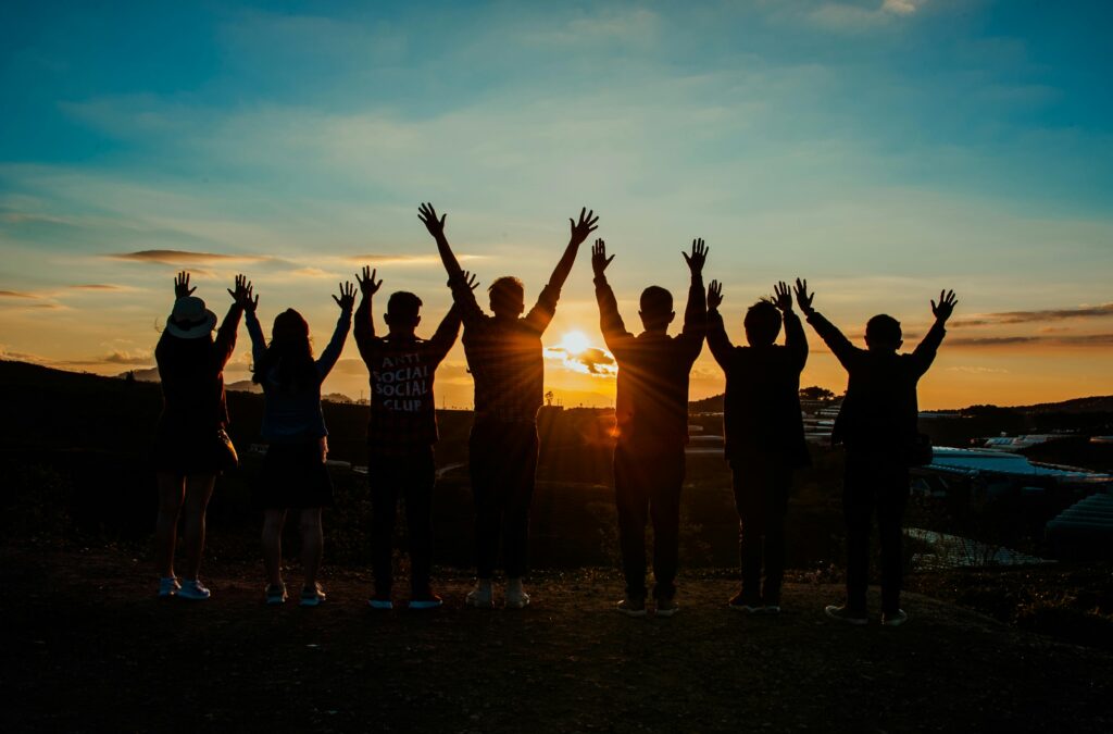 home A diverse group of friends raises their arms in celebration against a vibrant sunset backdrop.